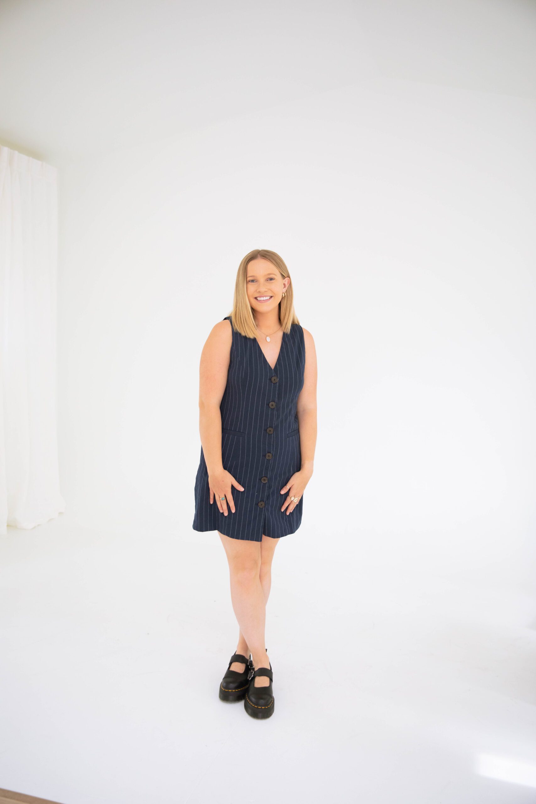 Friendly Denver therapist in a navy pinstripe dress standing and smiling in a bright, minimal studio setting