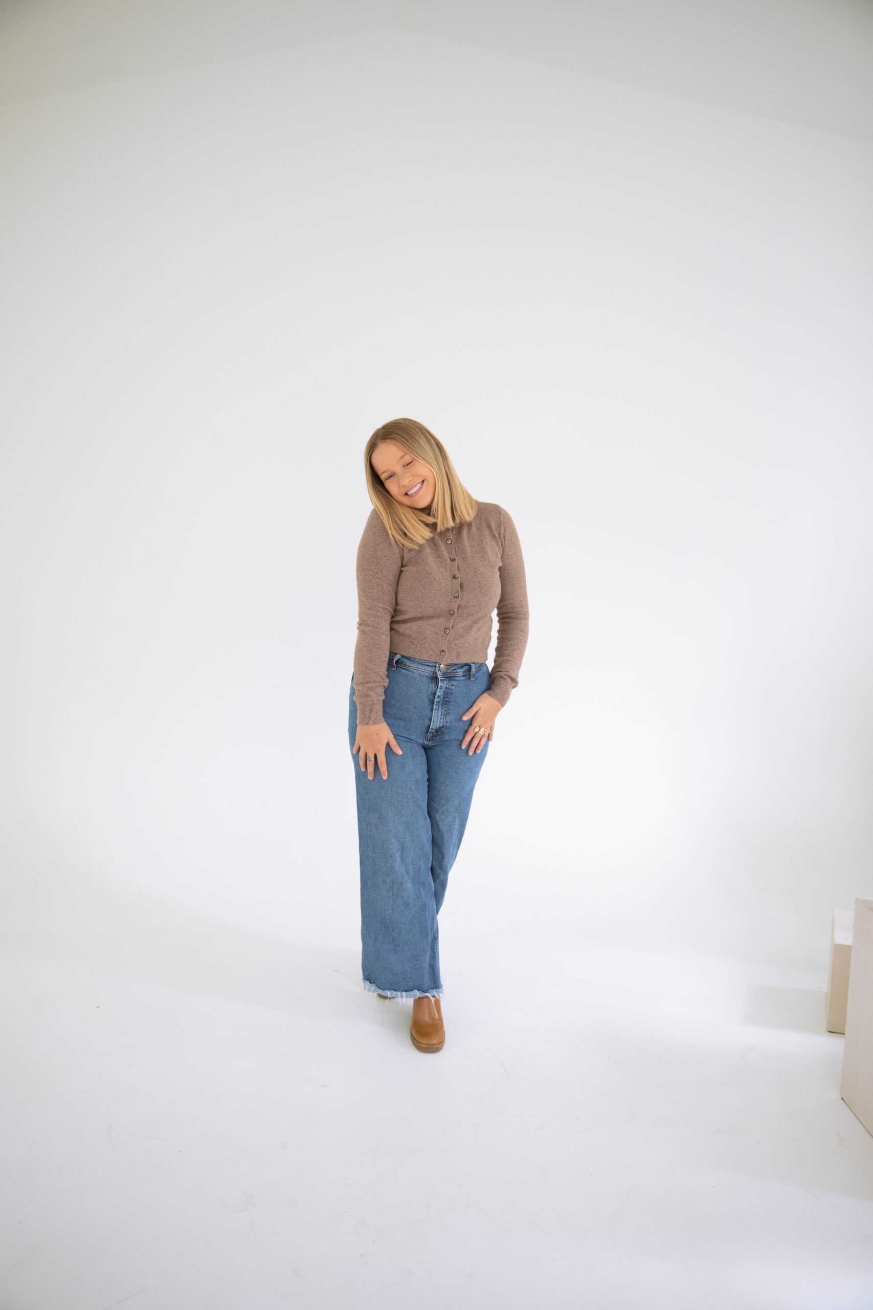 Woman in a brown sweater and wide-leg jeans standing in a studio, smiling with a relaxed, friendly posture.