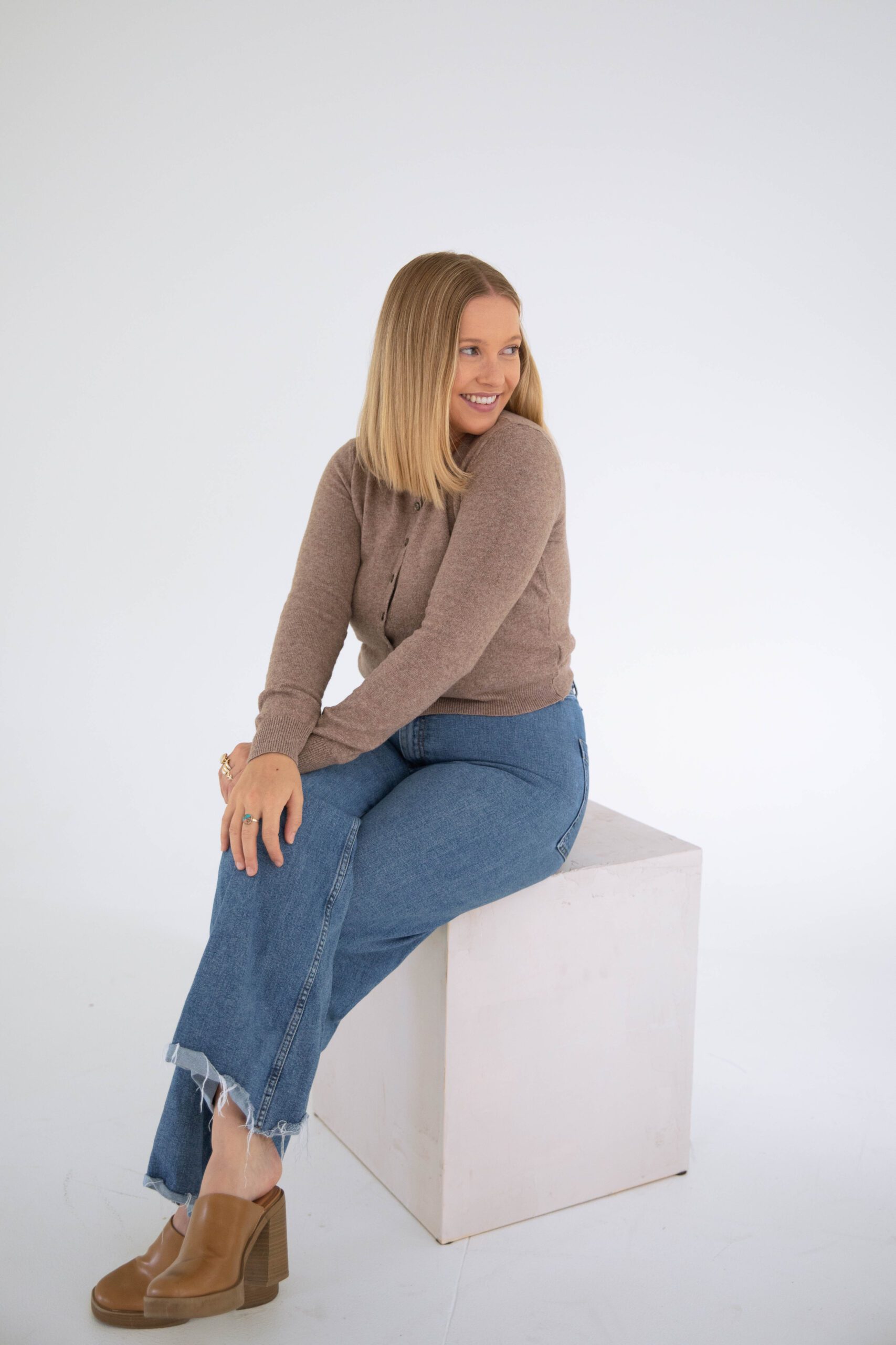 Woman sitting on a white cube in a studio, smiling and looking off to the side in a cozy brown sweater, jeans, and tan clogs.