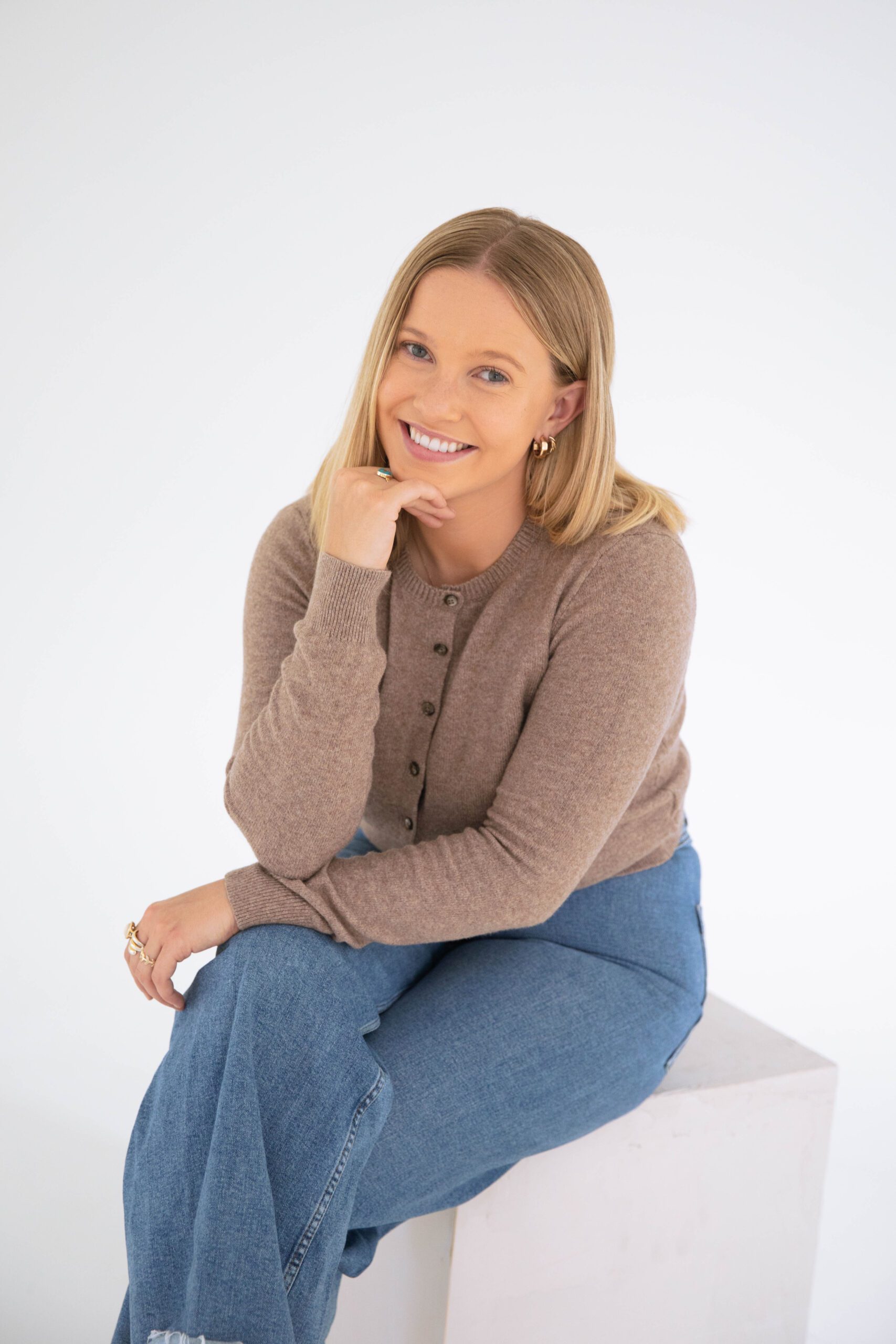 Close-up portrait of a smiling Denver therapist seated in a studio, resting her chin on her hand and wearing a neutral-toned sweater.