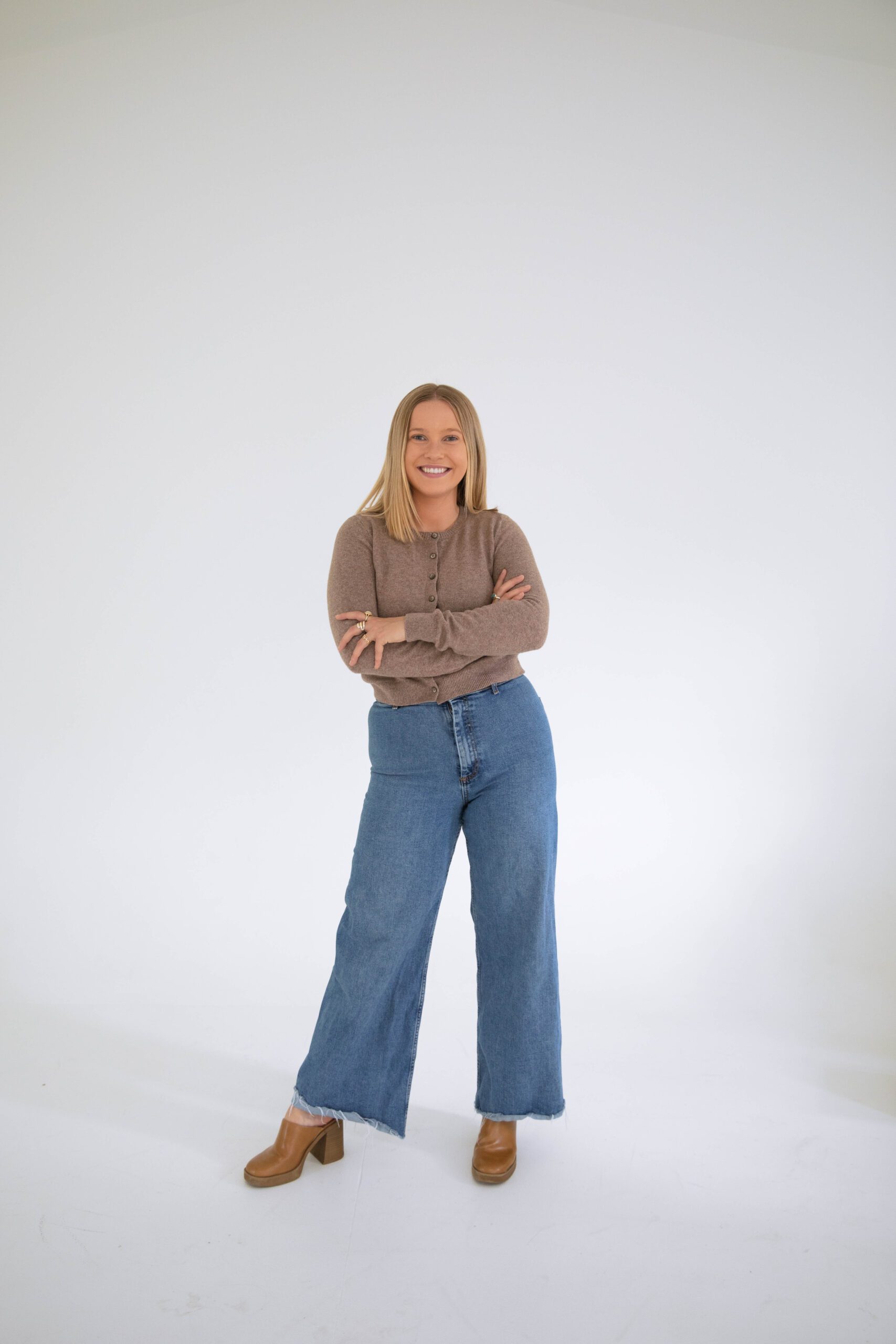 Confident Denver therapist standing with arms crossed, wearing a neutral cardigan and jeans in a modern white studio