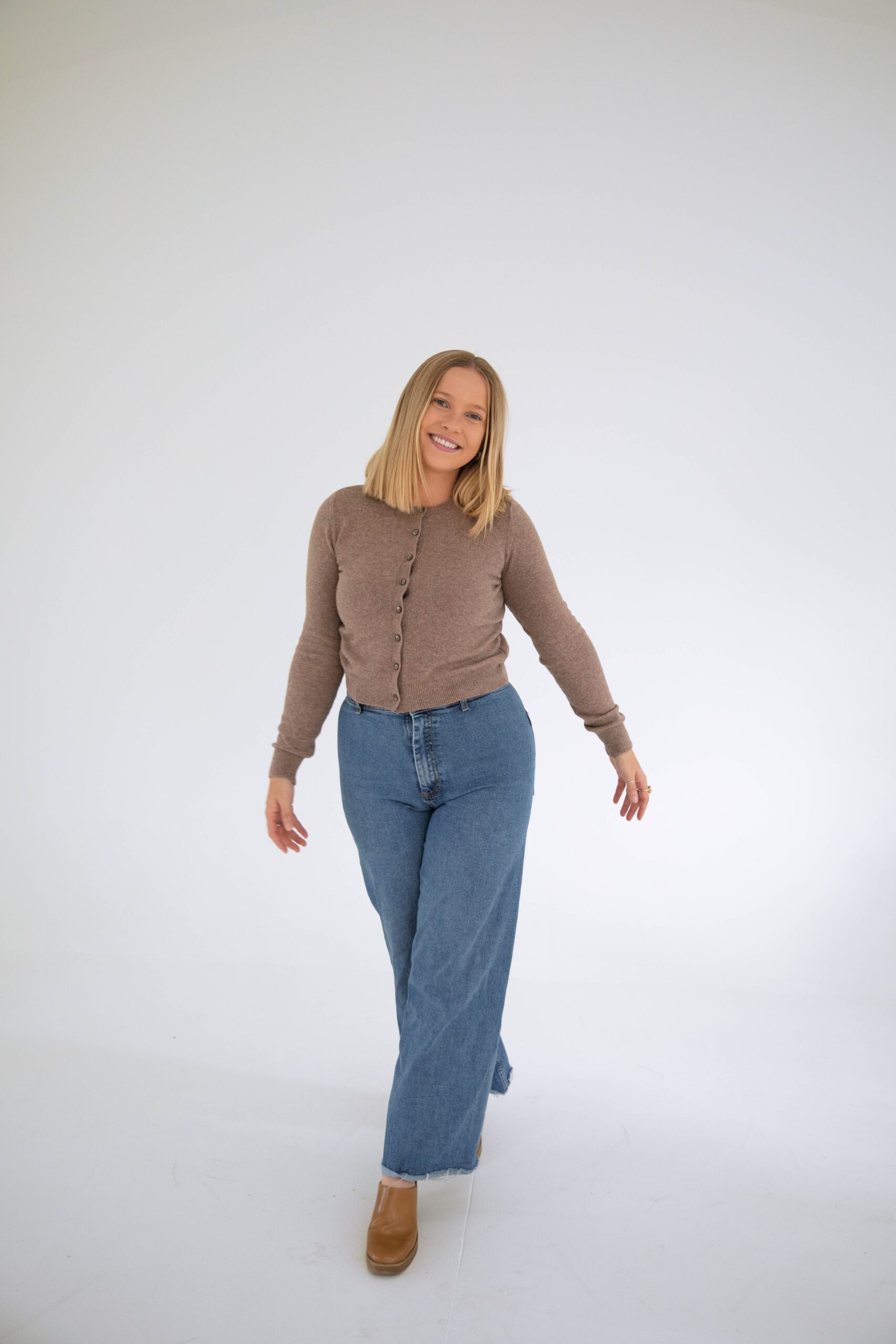 Casual portrait of a woman in a brown cardigan and wide-leg jeans walking forward in a light, airy studio
