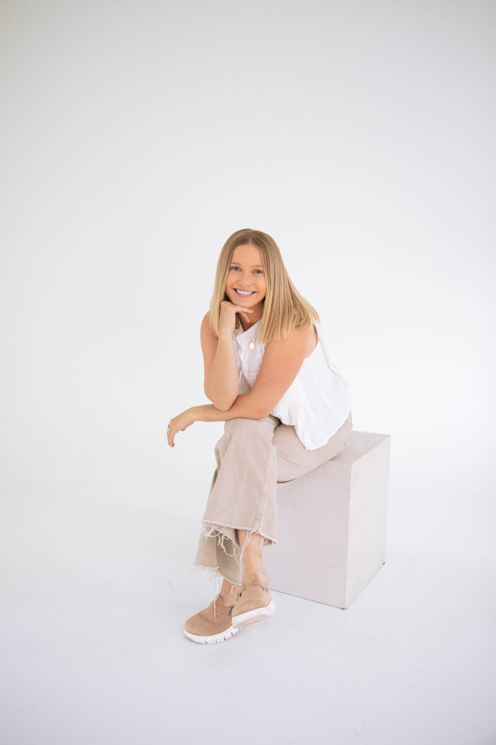 Warm and approachable EMDR therapist sitting on a white cube in a neutral studio, smiling with chin resting on hand
