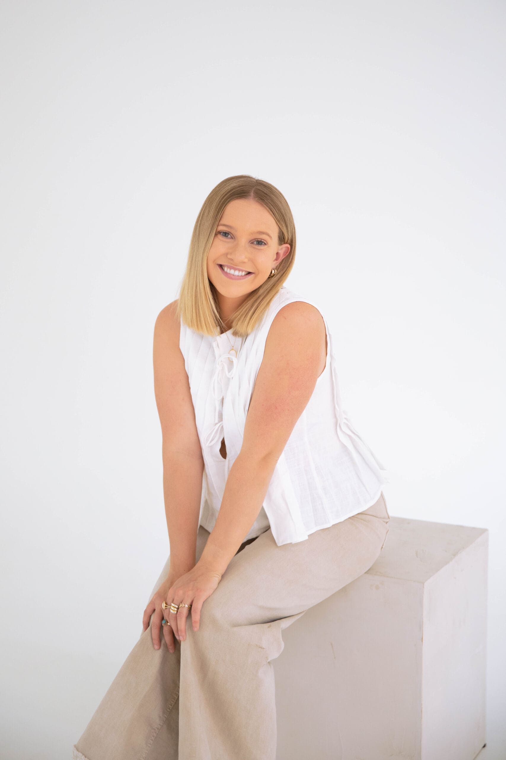 Friendly woman in a white blouse and neutral pants sitting and smiling in a bright, minimal studio setting