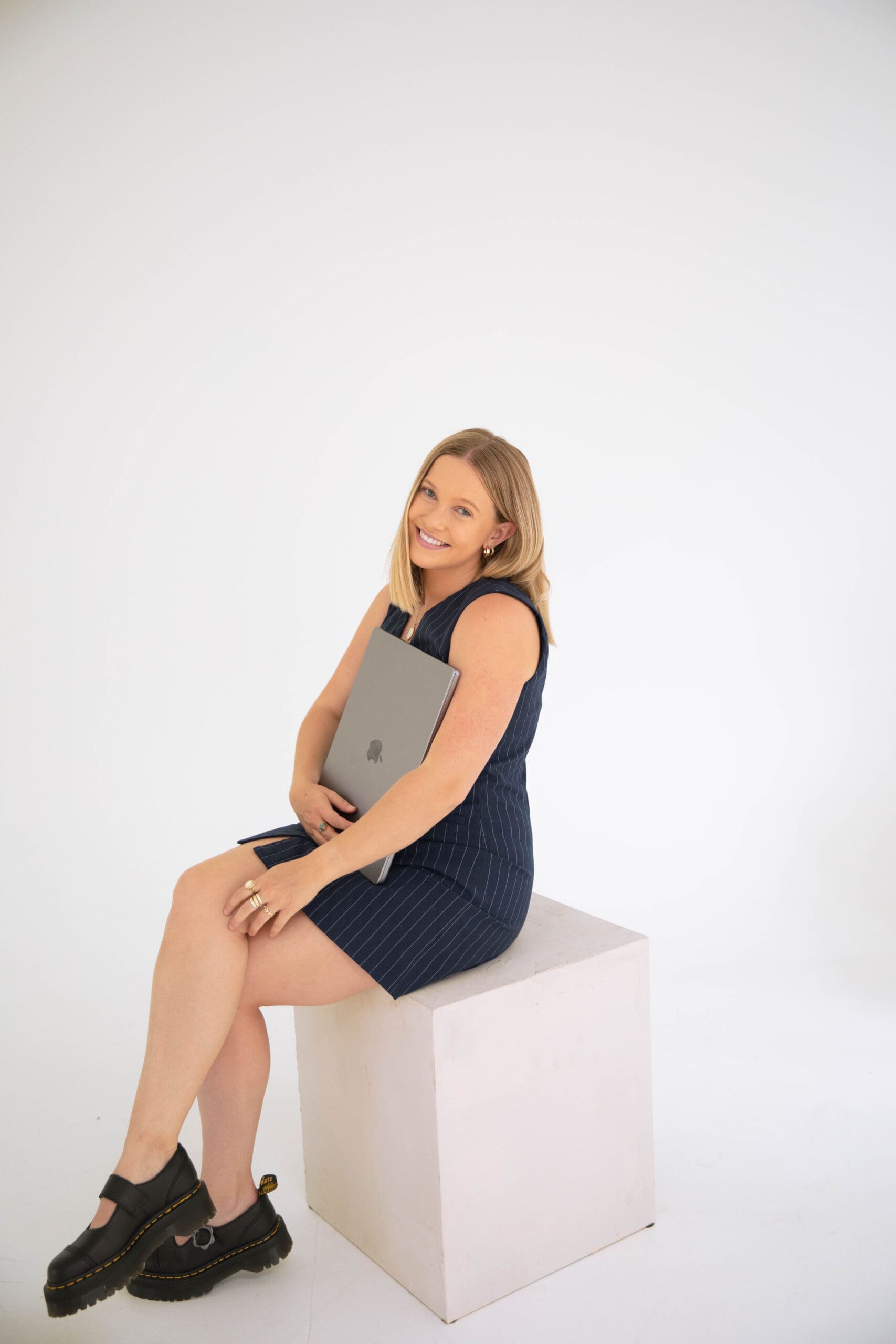 Confident EMDR therapist holding a laptop while seated on a white cube, smiling in a clean studio background

