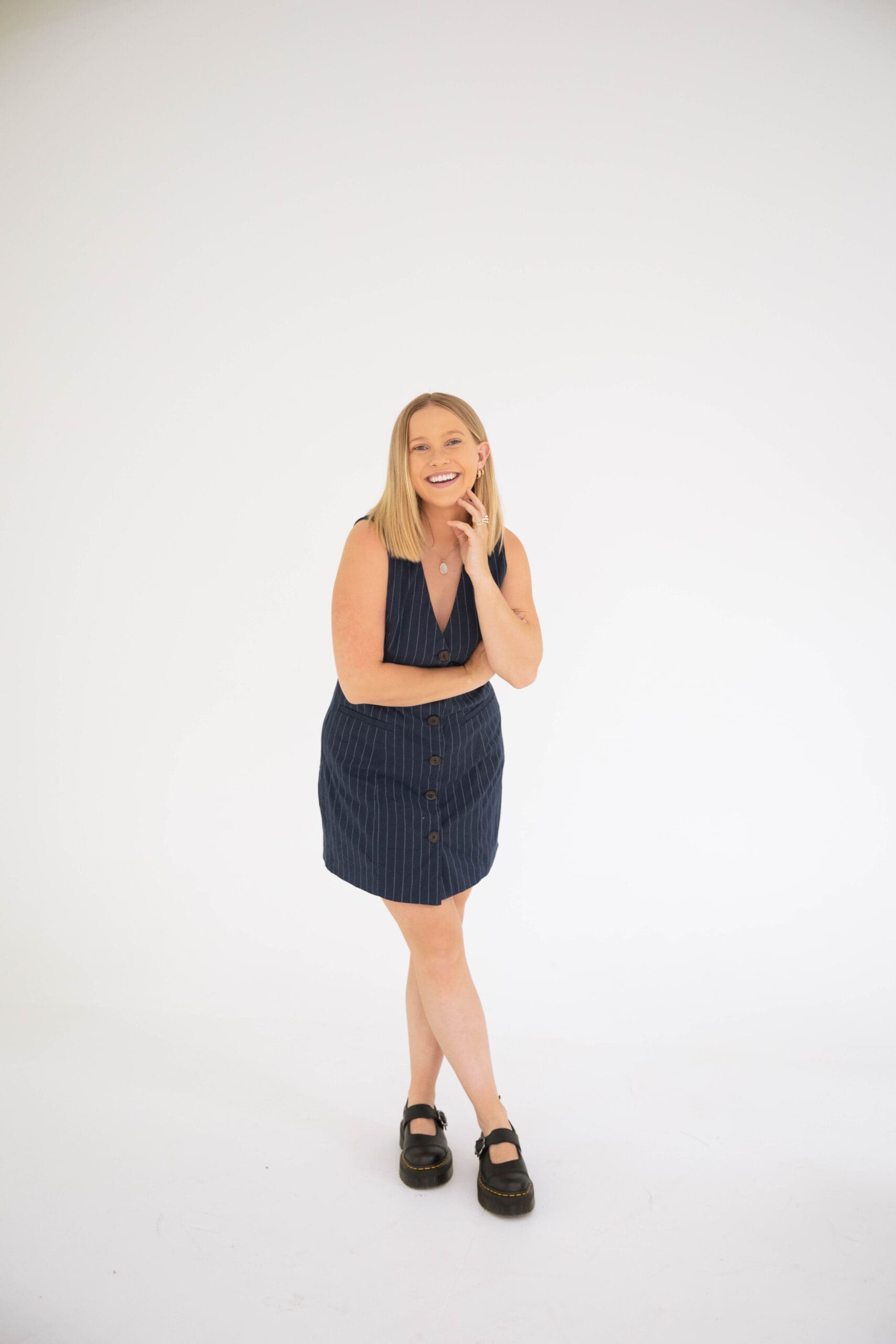 Woman in a navy pinstripe dress standing in a clean white studio with a relaxed and welcoming expression