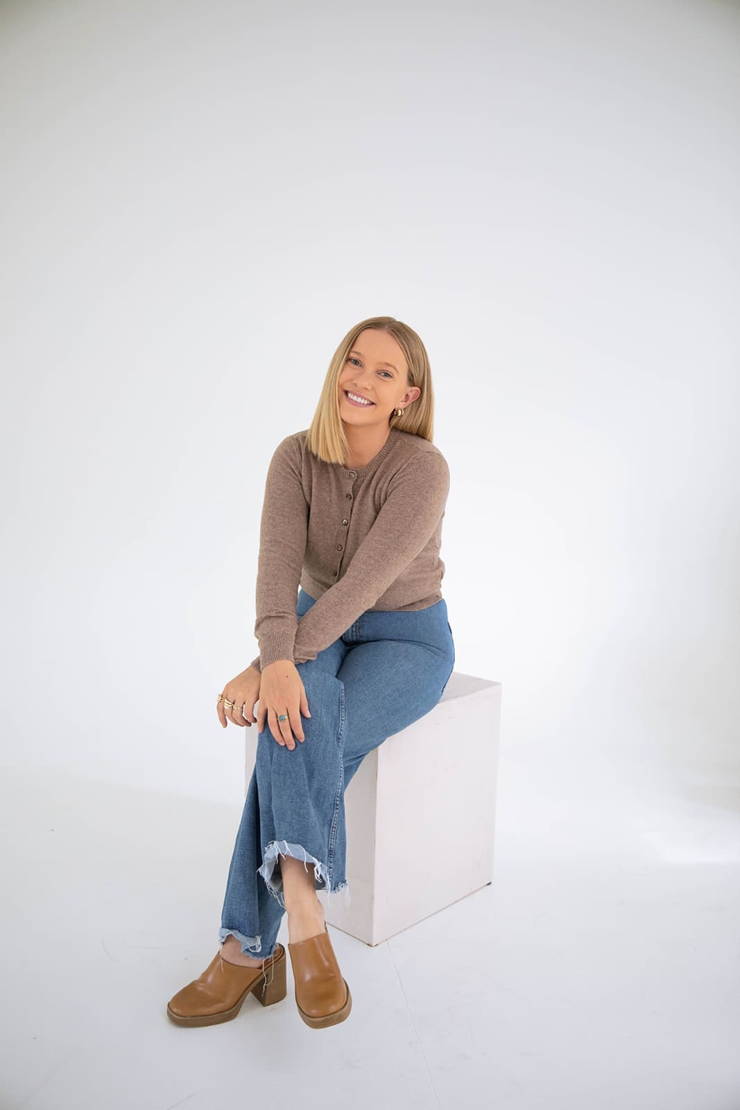 Denver therapist seated on a white studio block, smiling warmly in a relaxed and welcoming portrait.